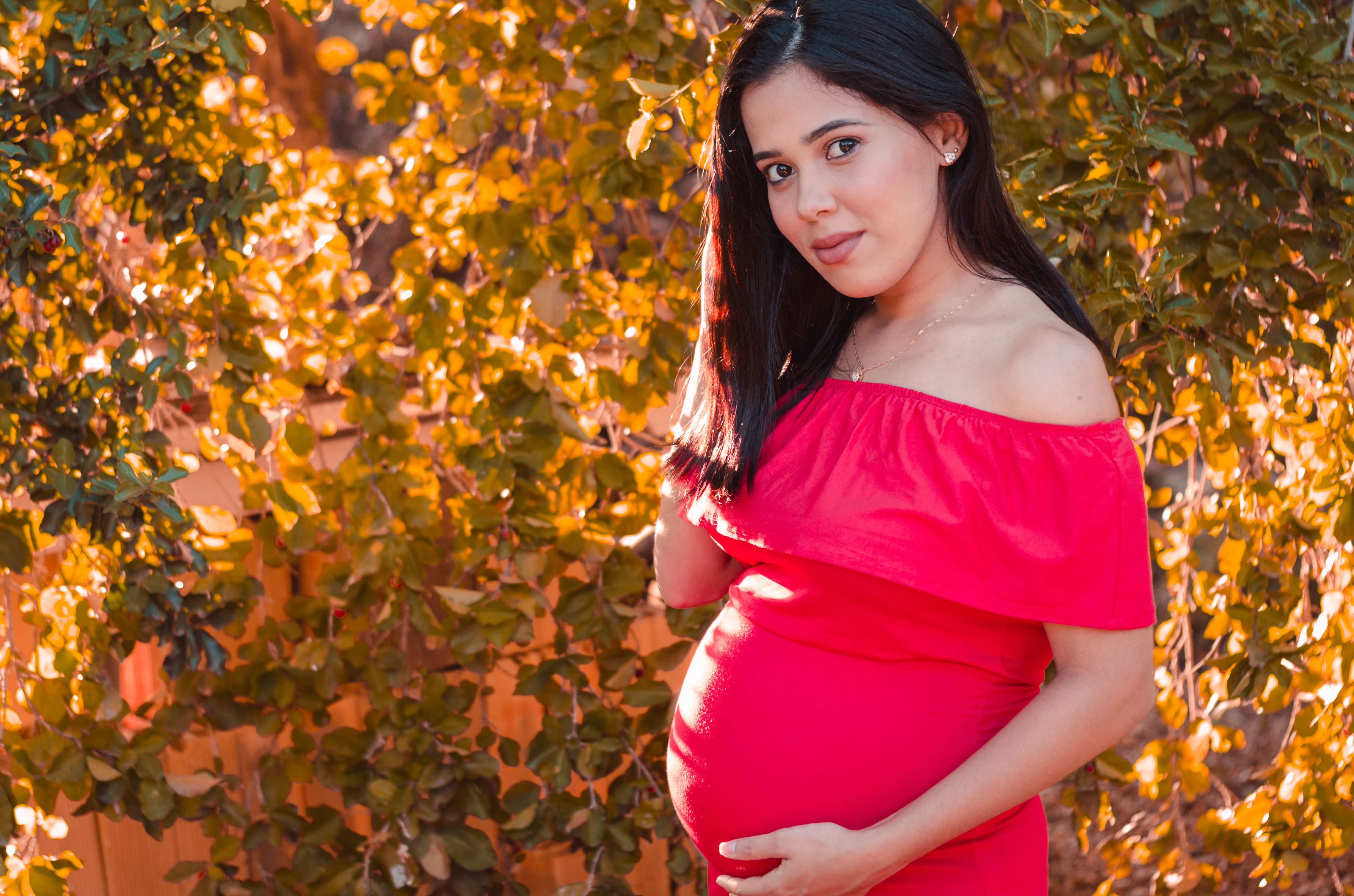 A pregnant woman in a vibrant red dress stands outdoors among golden autumn leaves, exuding warmth and beauty.