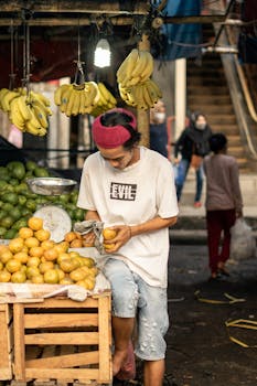 A fruit vendor arranges oranges at a busy market, surrounded by bananas and avocados.