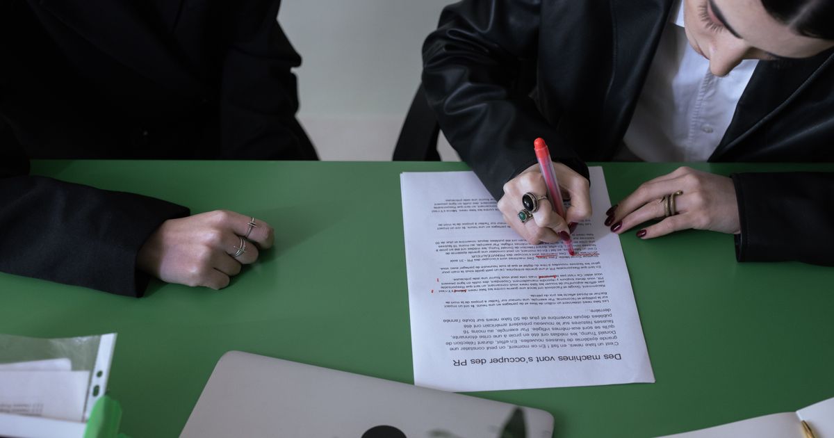Vue de dessus d'une personne qui corrige un texte imprimé au stylo rouge sur une table verte, un ordinateur portable fermé à côté d'elle.