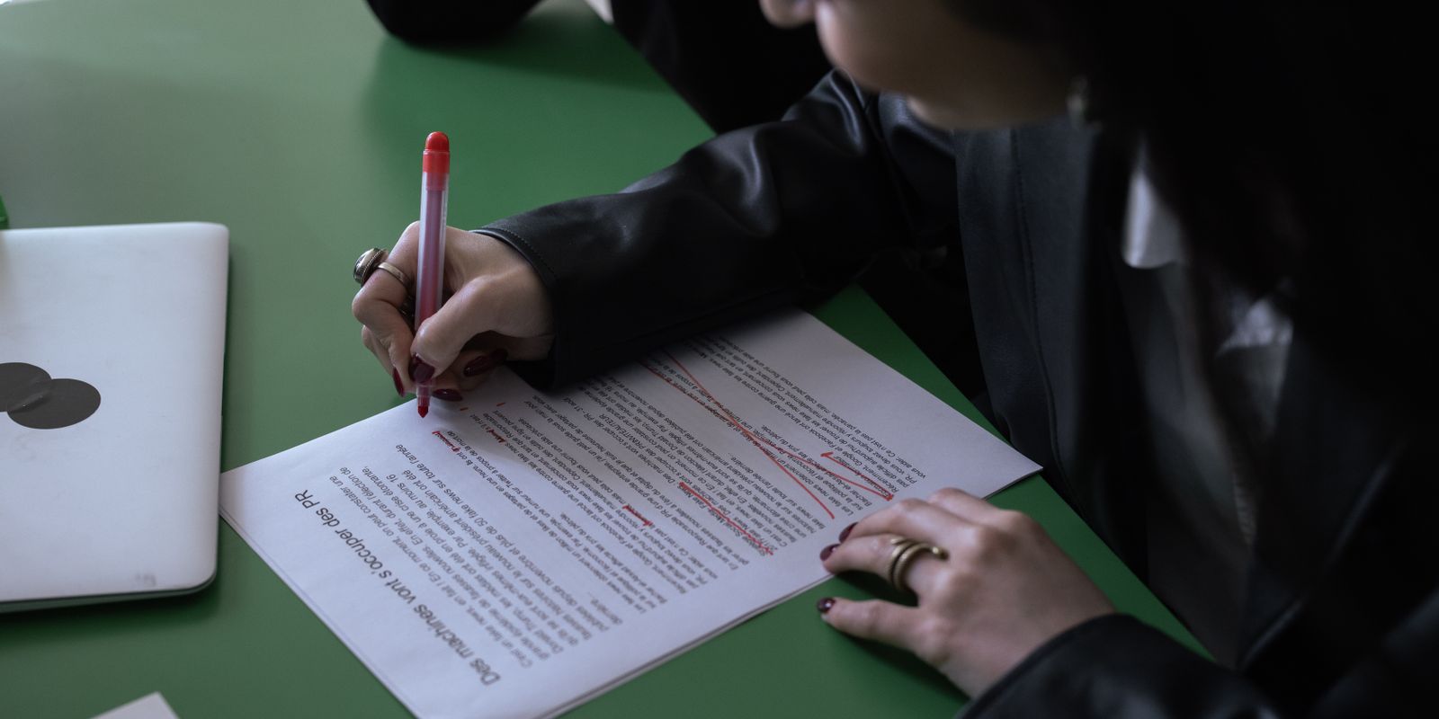 A woman in a suit uses a red pen to edit a printed document on a green table with a laptop.
