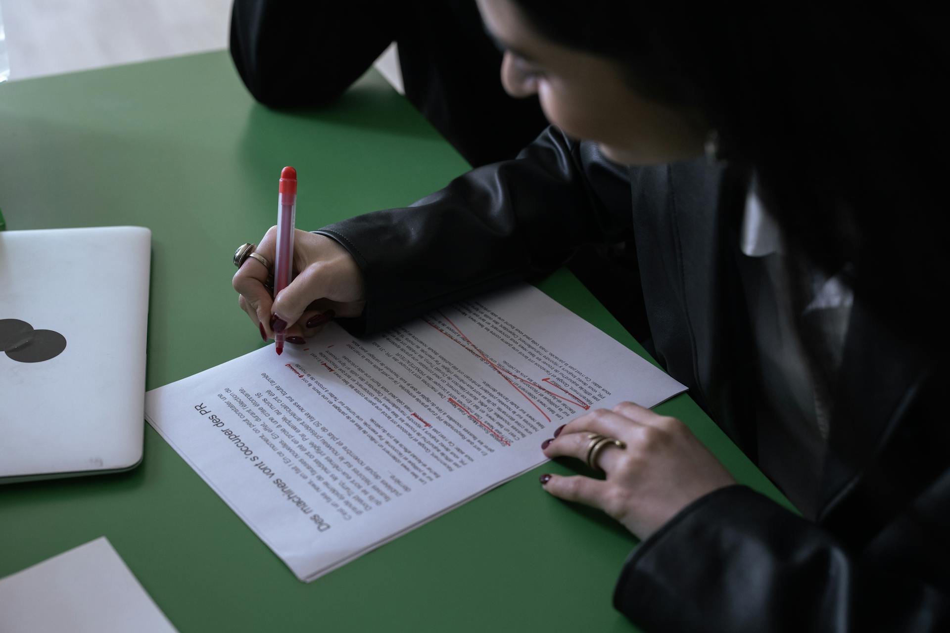 A woman in a suit uses a red pen to edit a printed document on a green table with a laptop.