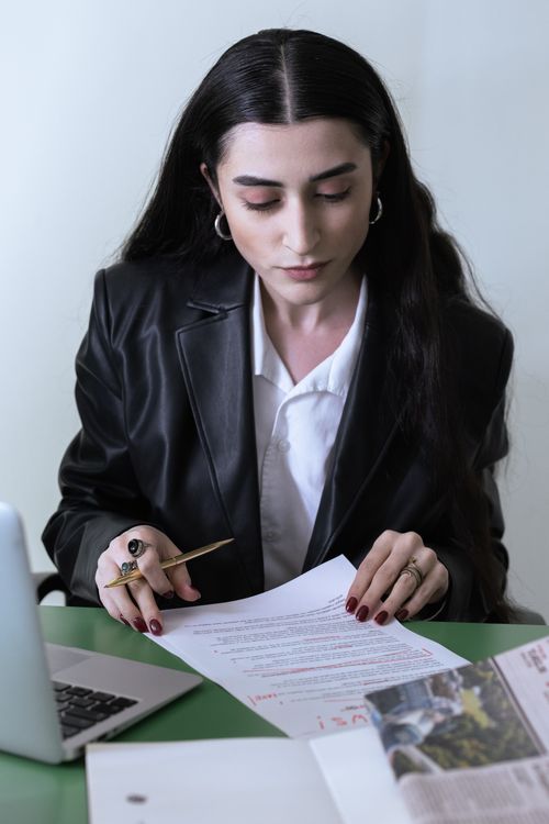 Free Professional woman in a leather coat reviewing documents next to a laptop in an office. Stock Photo
