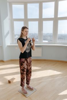 A woman meditates indoors on a sadhu board, holding tingsha bells with concentration.
