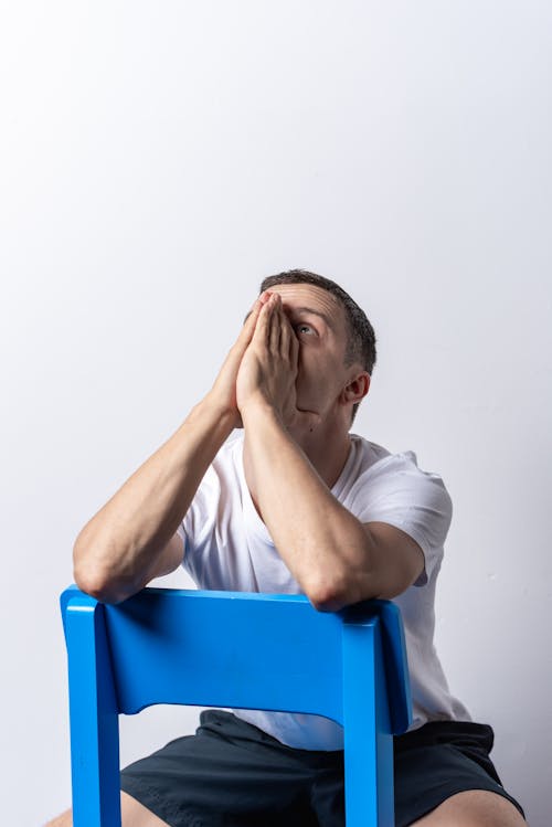 Man in White Shirt Sitting on Blue Chair Covering His Face With Hands and Looking Up 