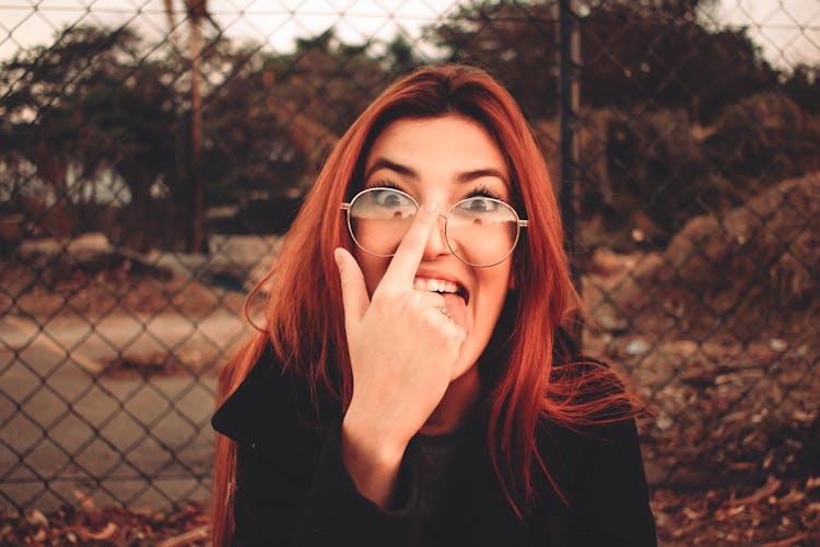 Funny Woman In Eyeglasses Touching Nose Against Grid Fence