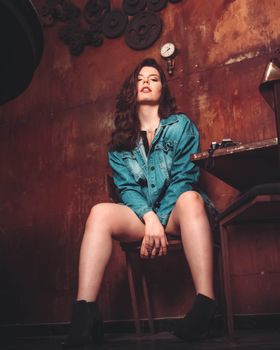 From below of young provocative female in boots sitting on chair while looking at camera against rusty wall in garage