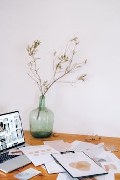 A clean workspace featuring a laptop, documents, and a decorative vase with branches.