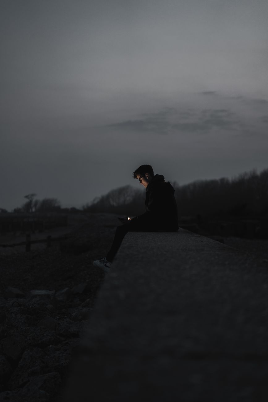 A solitary man sits alone at night, illuminated by phone light, in a dark outdoor setting.. solitary, sits, alone