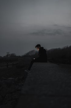 A solitary man sits alone at night, illuminated by phone light, in a dark outdoor setting.. solitary, sits, alone