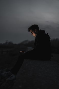A man sits outdoors in twilight, focused on his phone, creating a contemplative silhouette.