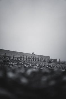 A solitary figure sits on a ledge overlooking a pebbly beach in this moody, black-and-white coastal scene.
