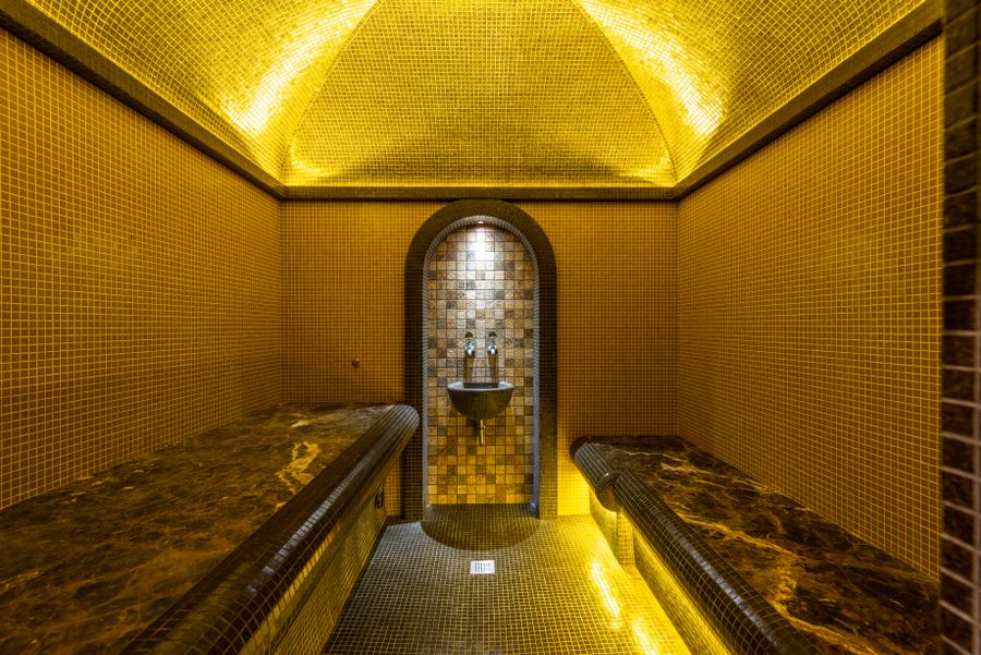 A hammam lined with gold mosaic under a softly lit vaulted ceiling, with heated marble benches
