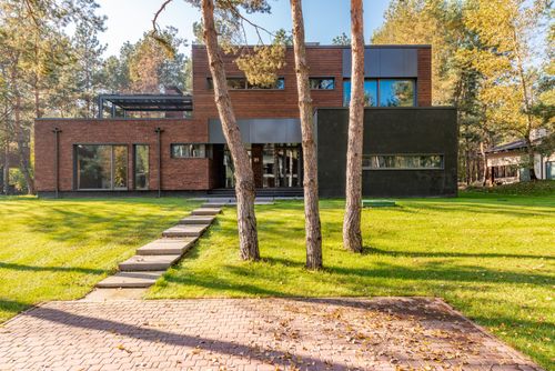 Free Contemporary house with large glass windows surrounded by lush greenery and a paved pathway. Stock Photo