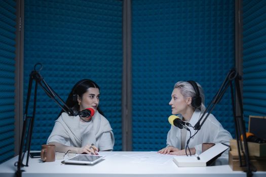 Two women in a soundproof studio recording a podcast with microphones and equipment.