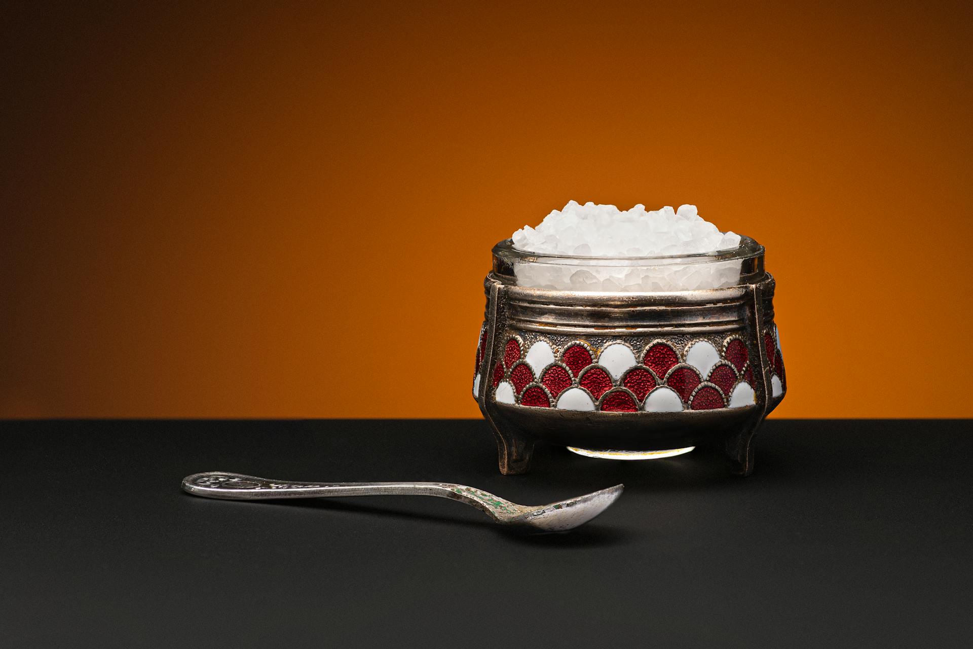 An ornate metal bowl filled with rock salt beside a decorative spoon on a black surface with a warm background.
