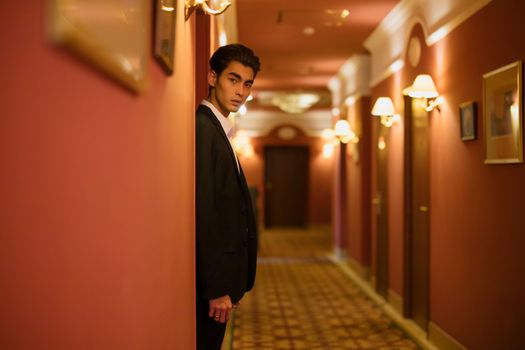 A fashionable man in a sleek black suit poses confidently in an elegant hotel hallway.