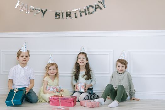 Four cheerful kids celebrating a birthday with gifts and party hats indoors.
