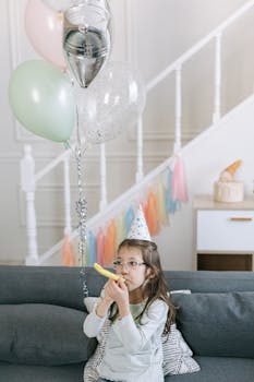 A young girl wearing a party hat blowing a whistle in a festive birthday setting indoors.