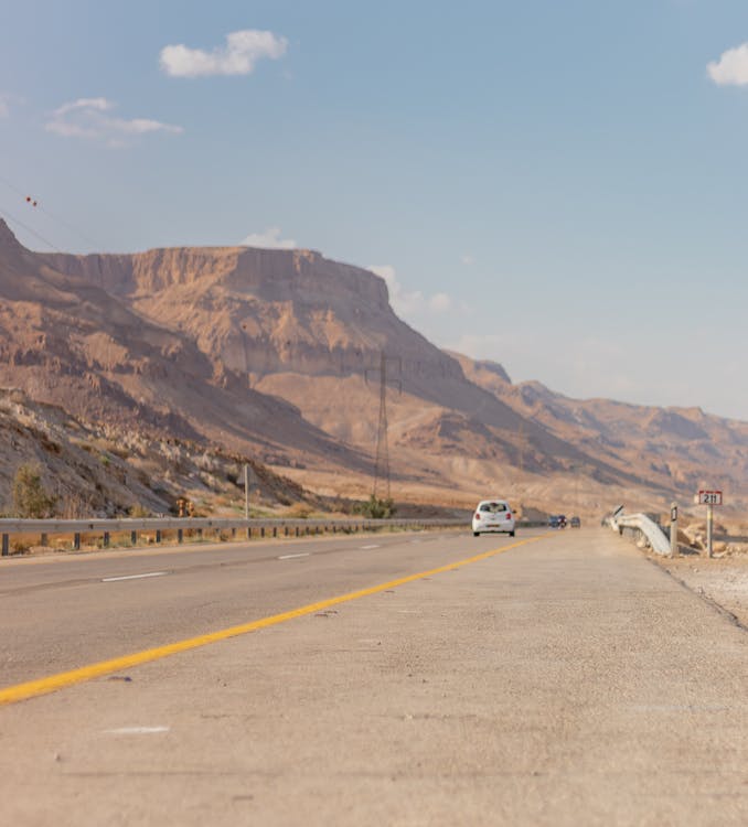 Straight asphalt roadway running along rocky terrain with car driving in distance under blue sky on sunny day