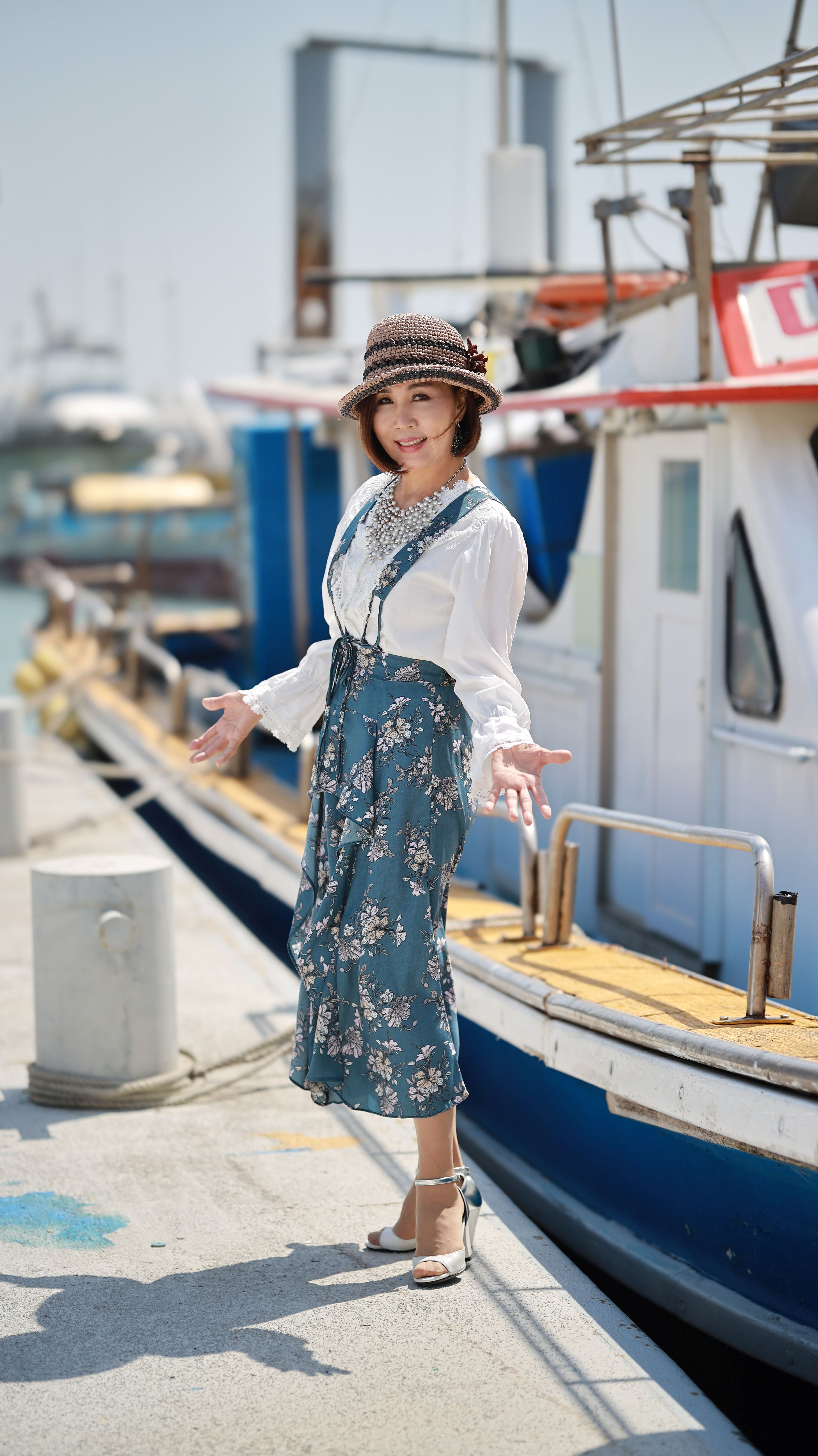 Smiling woman in floral dress and hat, posing by boats at the harbor on a sunny day.