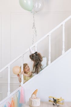 Two girls joyfully celebrating a birthday with balloons and cake on a staircase.
