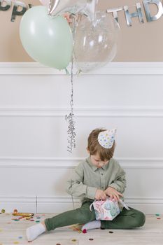 Young boy opening a present at a birthday party with balloons and confetti indoors.