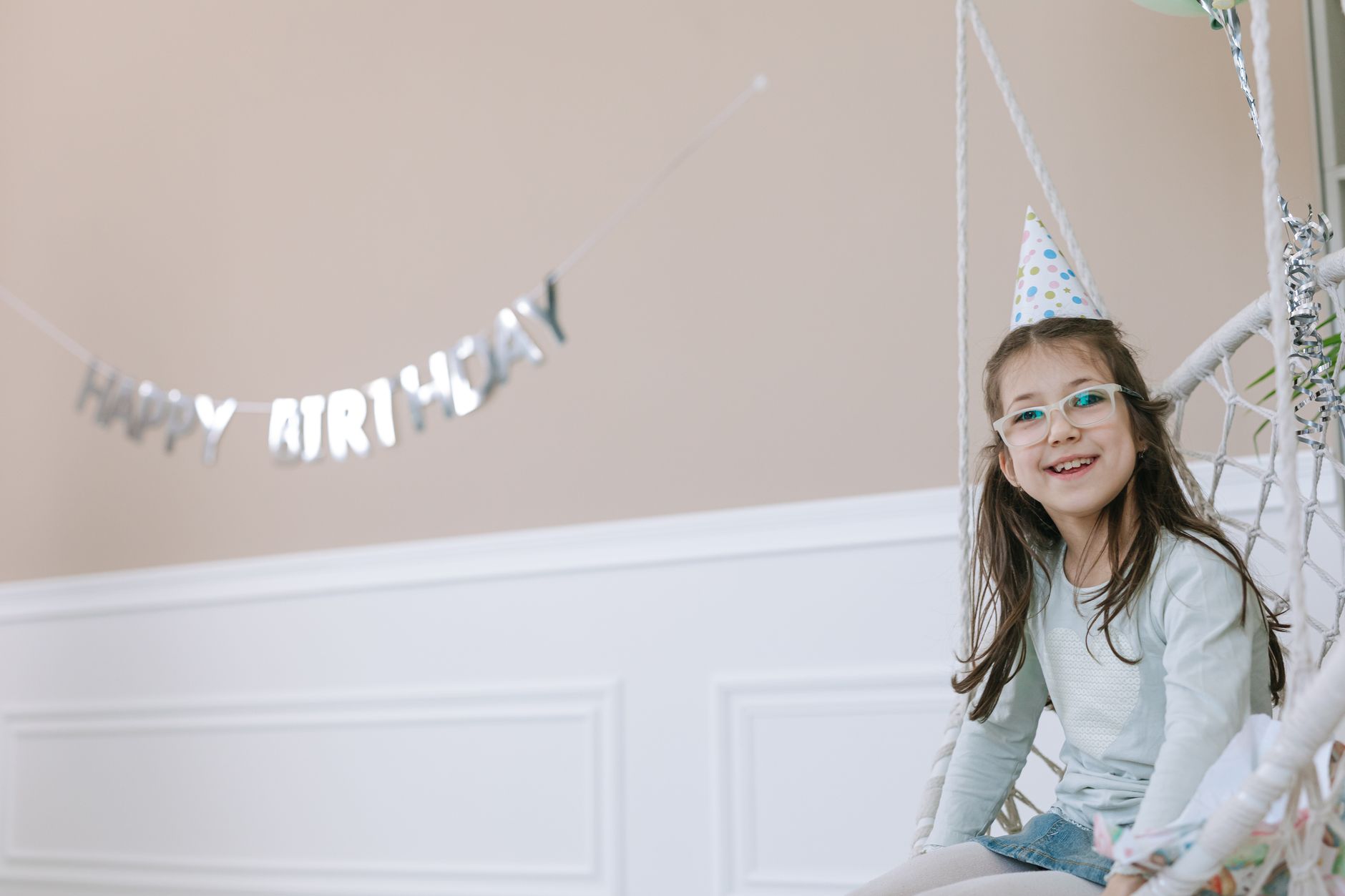 best birthday party venues in cairo for kids — Young girl wearing glasses and party hat smiling on a swing indoors.
