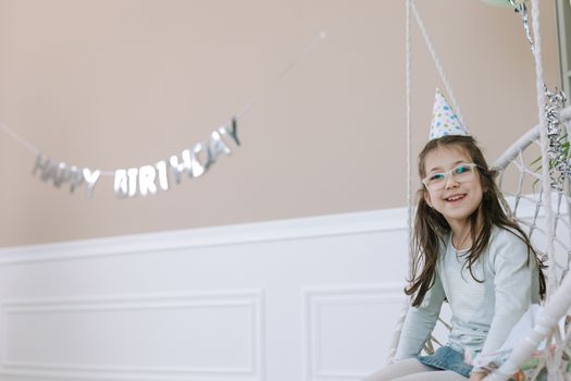 Young girl wearing glasses and party hat smiling on a swing indoors.