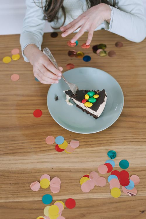 Free A child is eating a slice of cake topped with colorful candies, surrounded by confetti, on a wooden table. Stock Photo