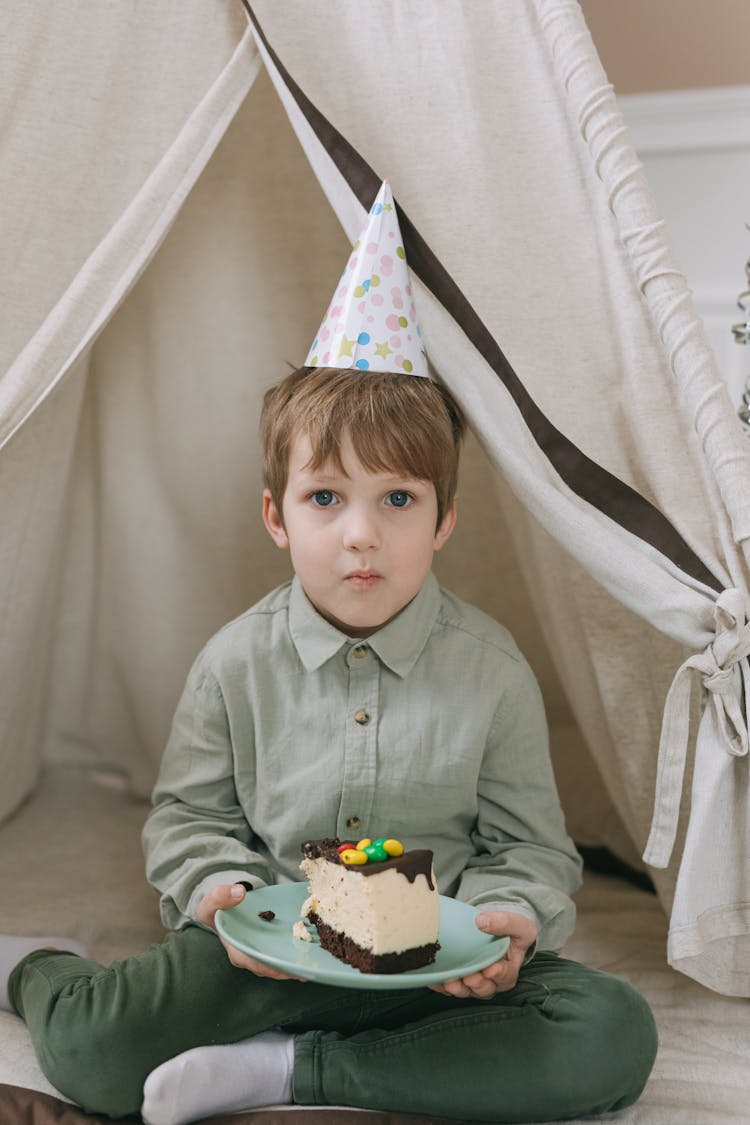 A Boy With A Birthday Hat Sitting In A Tent With A Slice Of Cake On Plate