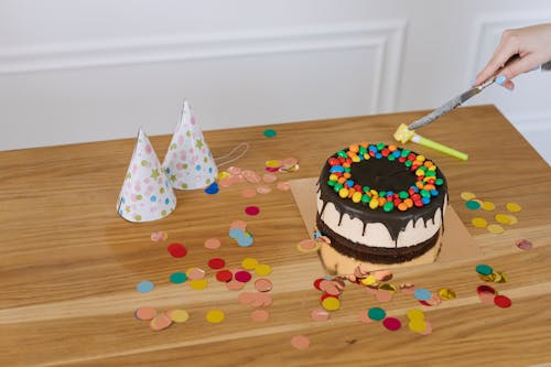 A Person's Hand Slicing a Cake Near Party Hats
