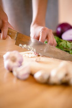 Person chopping garlic with a knife on a wooden cutting board indoors.