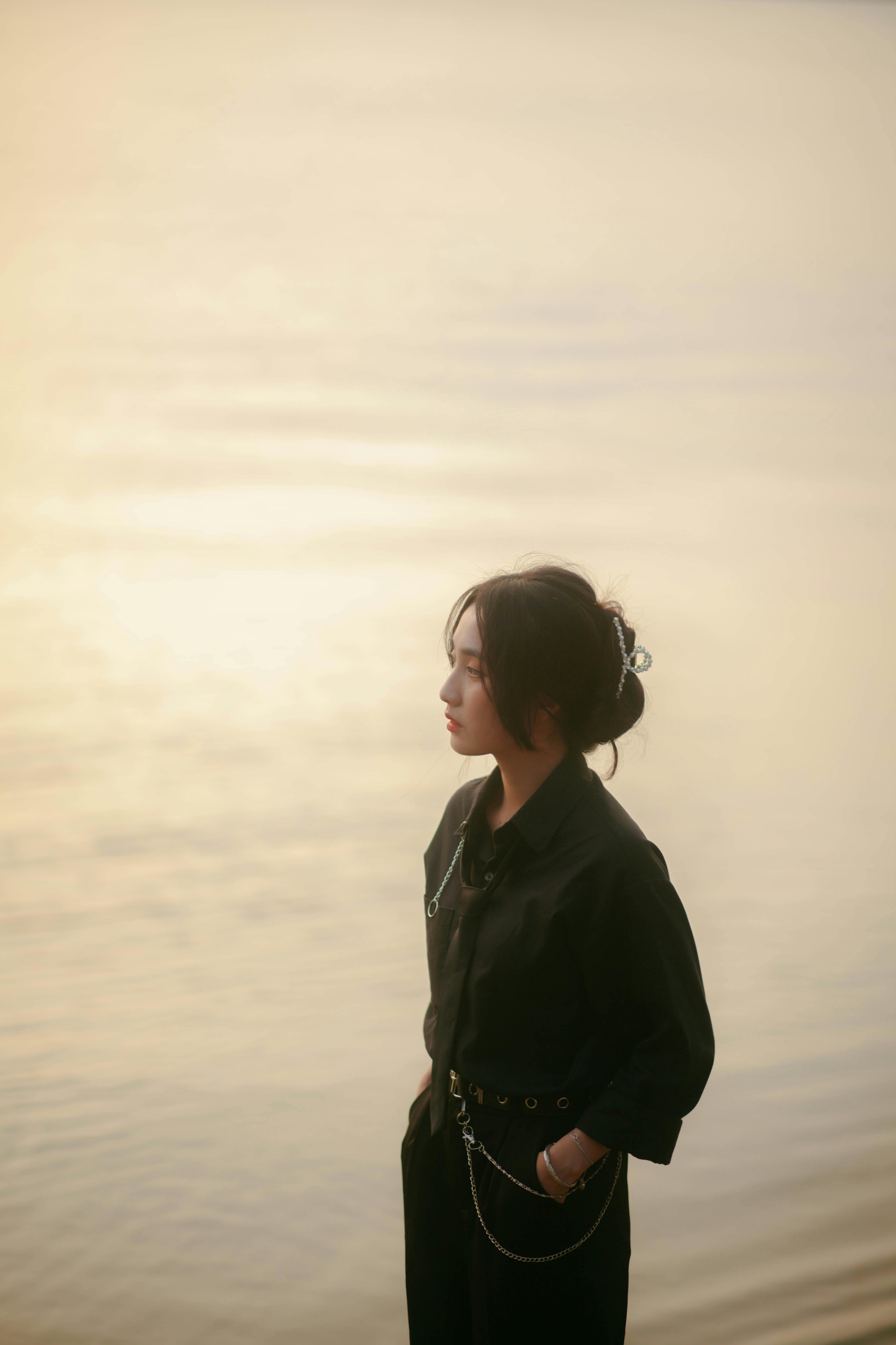 A woman dressed in black poses elegantly by serene water during sunset, capturing a moment of quiet reflection.