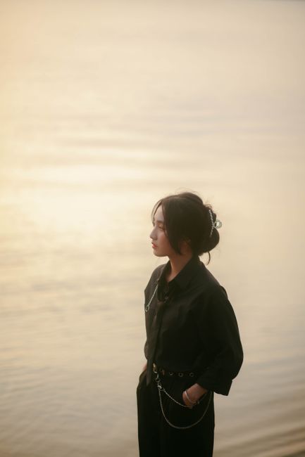 A woman dressed in black poses elegantly by serene water during sunset, capturing a moment of quiet reflection.