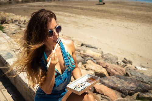 Free A woman with long hair and sunglasses eats sushi with chopsticks by the beach. Stock Photo
