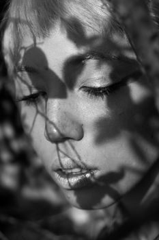 Black and white of crop young tender female with foliage shades on face looking down in sunlight