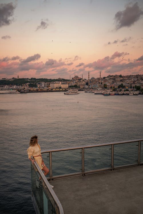 Free Distant anonymous female tourist admiring rippling sea while standing in harbor against moored ship in coastal town at sunset time Stock Photo