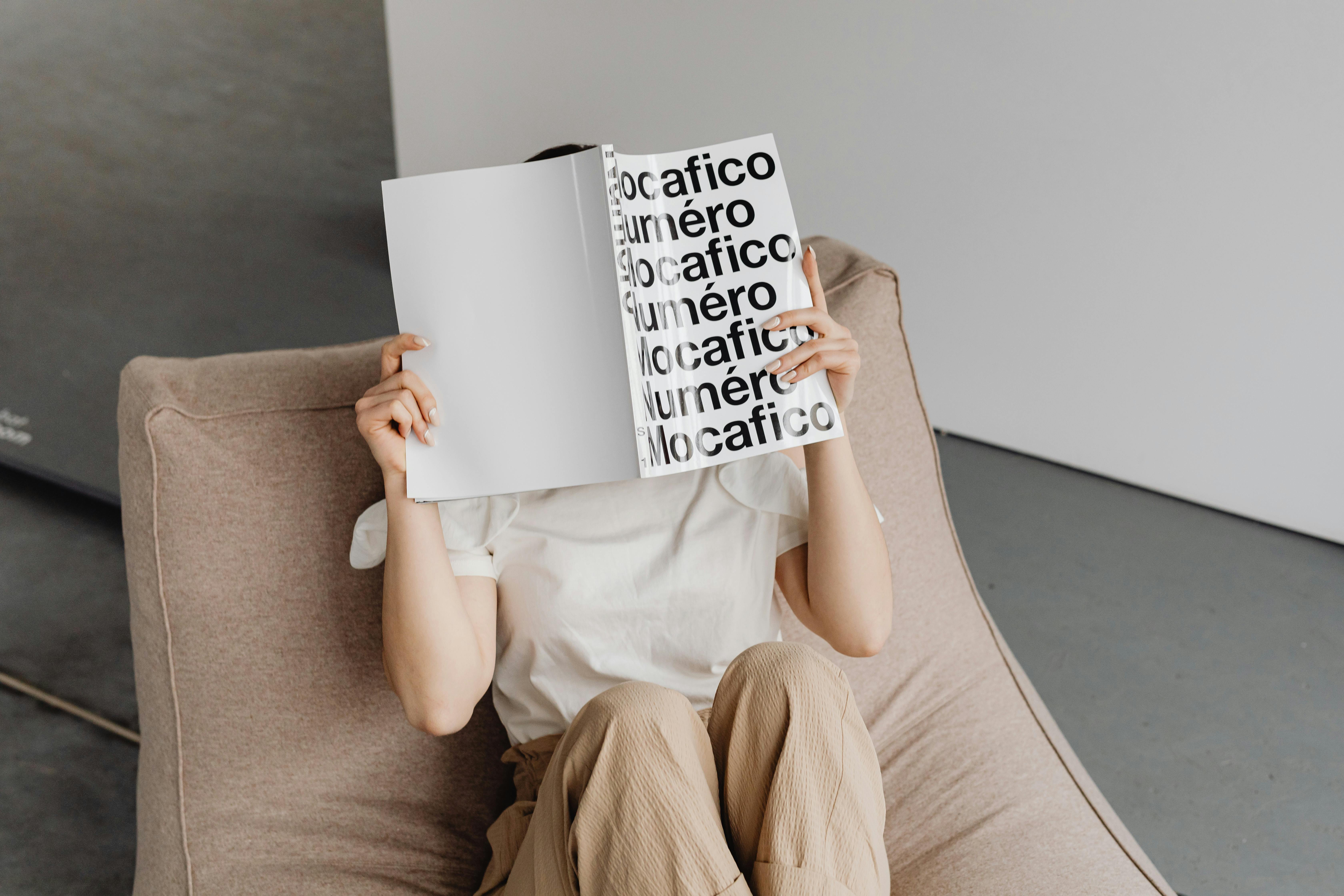 An individual comfortably sits in a modern setting, holding a book with their face obscured.