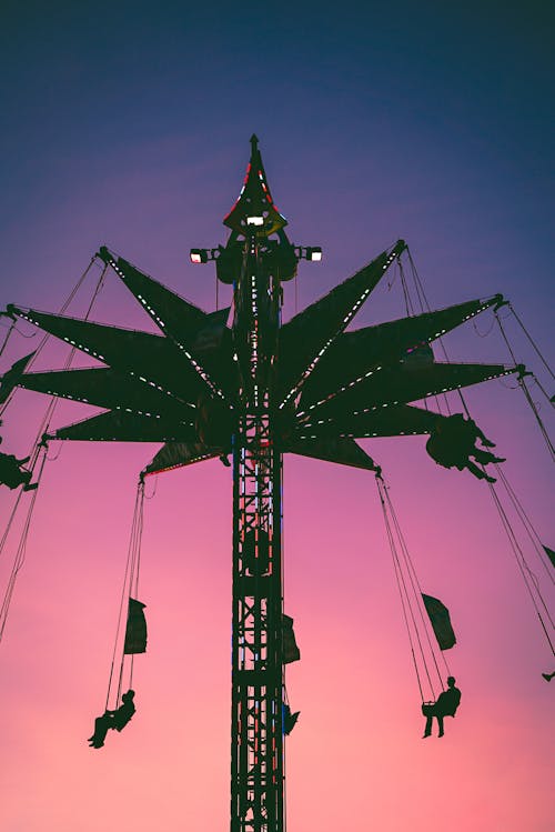 From below silhouette of unrecognizable people riding Chair O Planes against cloudless bright pink and purple sunset sky in amusement park