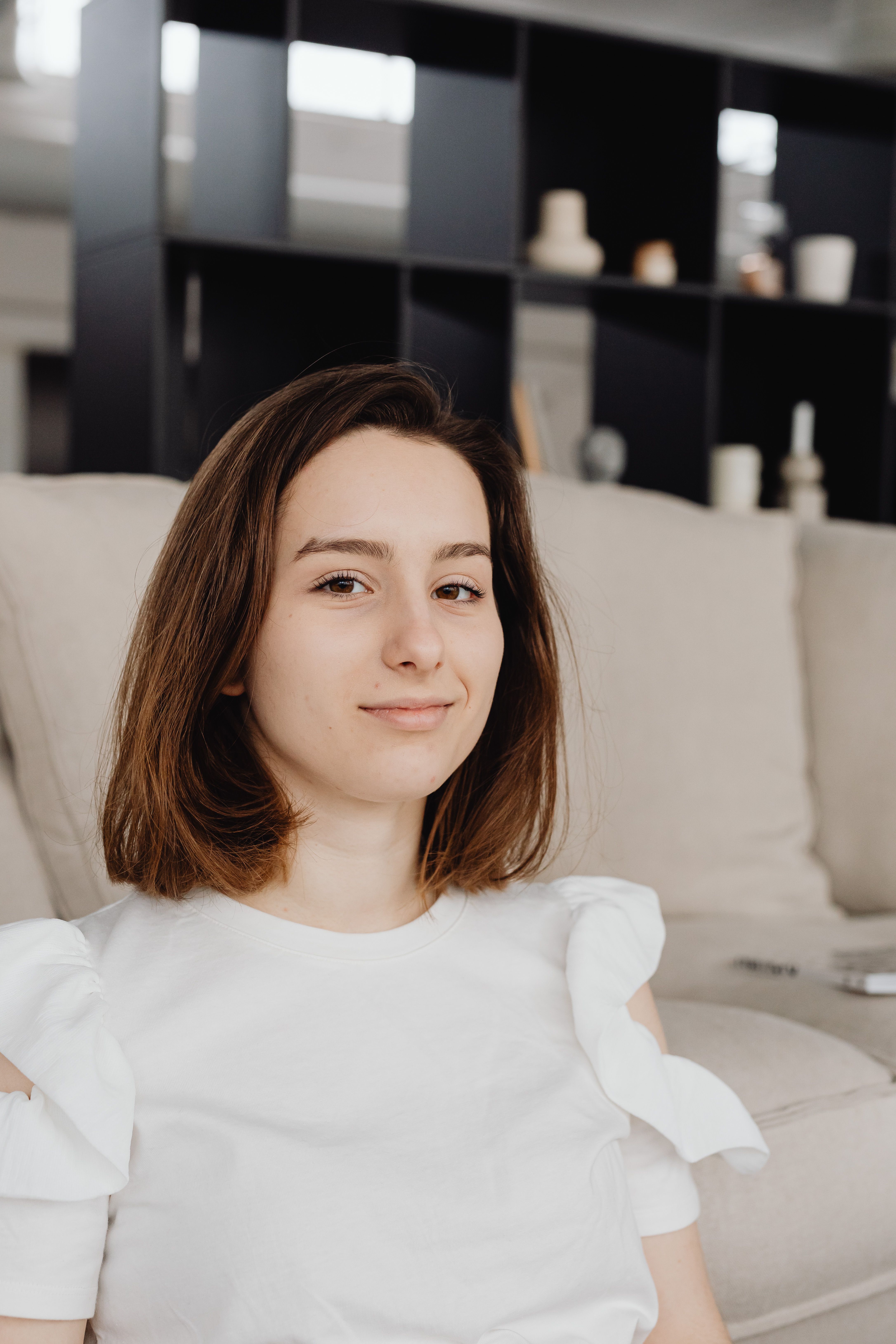Portrait of a young woman sitting on a sofa indoors, smiling gently with a bookshelf in the background.