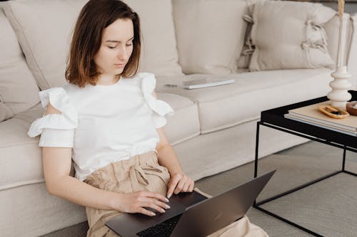Free Woman in White Shirt Sitting on the Floor beside Beige Sofa while using Laptop Stock Photo