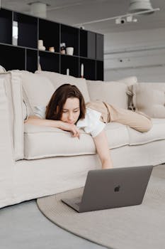Woman lying on a sofa working remotely from home on a laptop computer.