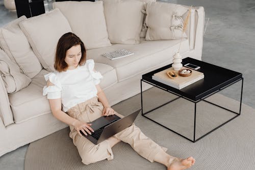 Woman in White Shirt Sitting on Brown Rug beside Beige Sofa while Using Laptop