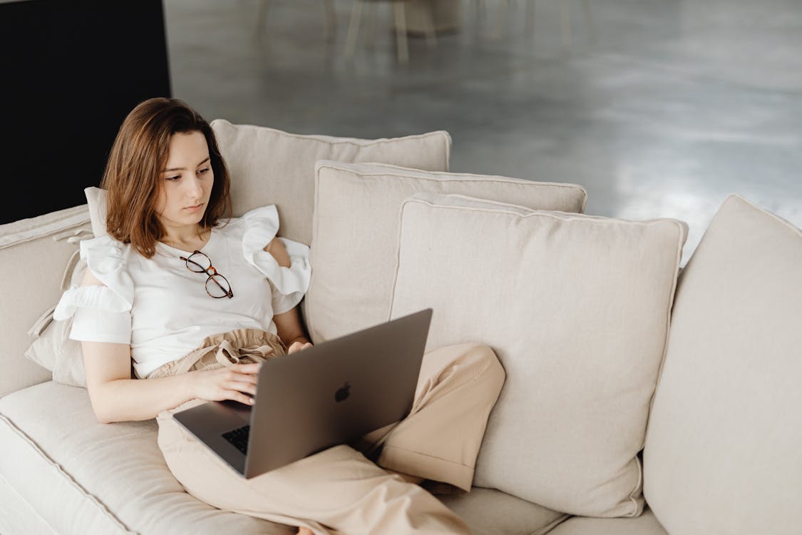 Free Woman in casual attire working on a laptop while lounging on a couch, emphasizing remote work. Stock Photo