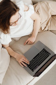 Overhead view of a woman using a laptop while lying on a couch, enjoying a relaxed home environment.