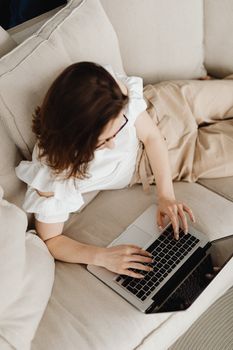 Woman comfortably using a laptop on a beige sofa indoors, viewed from above.