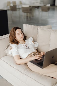 Woman lying on a couch working remotely on a laptop, embodying a comfortable home office setting.