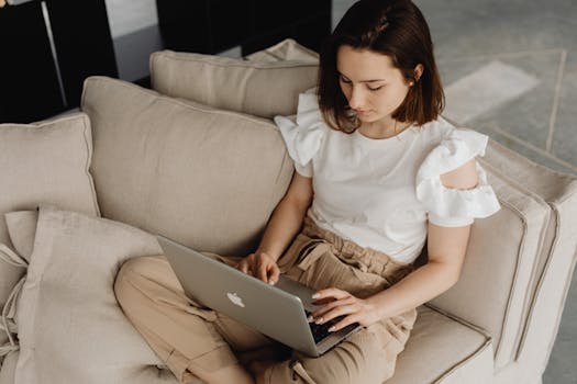 Young woman working on a laptop while sitting comfortably on a beige sofa indoors.