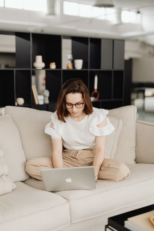Free Young woman wearing glasses working on laptop while seated on a sofa indoors, showcasing a modern lifestyle. Stock Photo