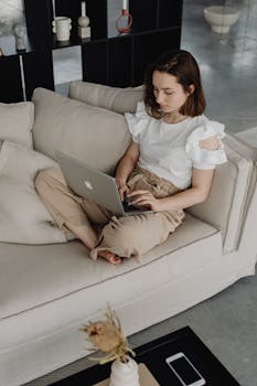 A young woman sits comfortably on a sofa using a laptop, capturing a cozy work-from-home setting.